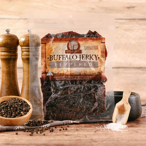 Packaged buffalo jerky with pepper, flanked by pepper mills and scattering of whole peppercorns on wooden surface.