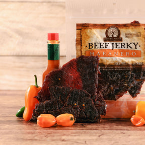 Habanero beef jerky displayed with fresh peppers and a sauce bottle on a wooden surface.