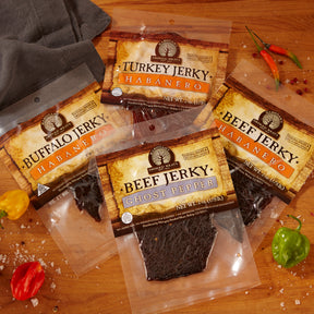 Variety of spicy meat jerky packages on a wooden surface with chili peppers and sea salt scattered around.